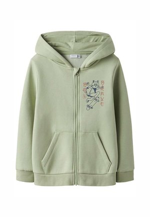 Sweat à capuche zippé vert clair avec poches avant et un dessin de chat en dessin animé faisant du skateboard sur la poitrine, avec un texte à côté.