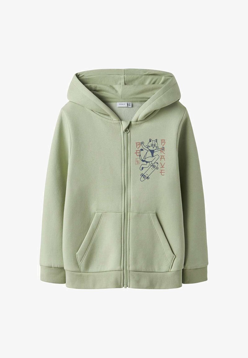 Sweat à capuche zippé vert clair avec poches avant et un dessin de chat en dessin animé faisant du skateboard sur la poitrine, avec un texte à côté.