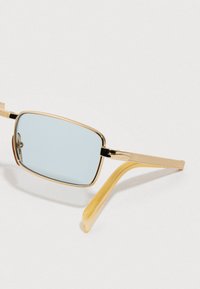 Pravougaone sunčane naočale sa zlatnim metalnim okvirom, svijetloplavim sočivima i prozirnim žutima vrhovima okvira na svijetloj pozadini.