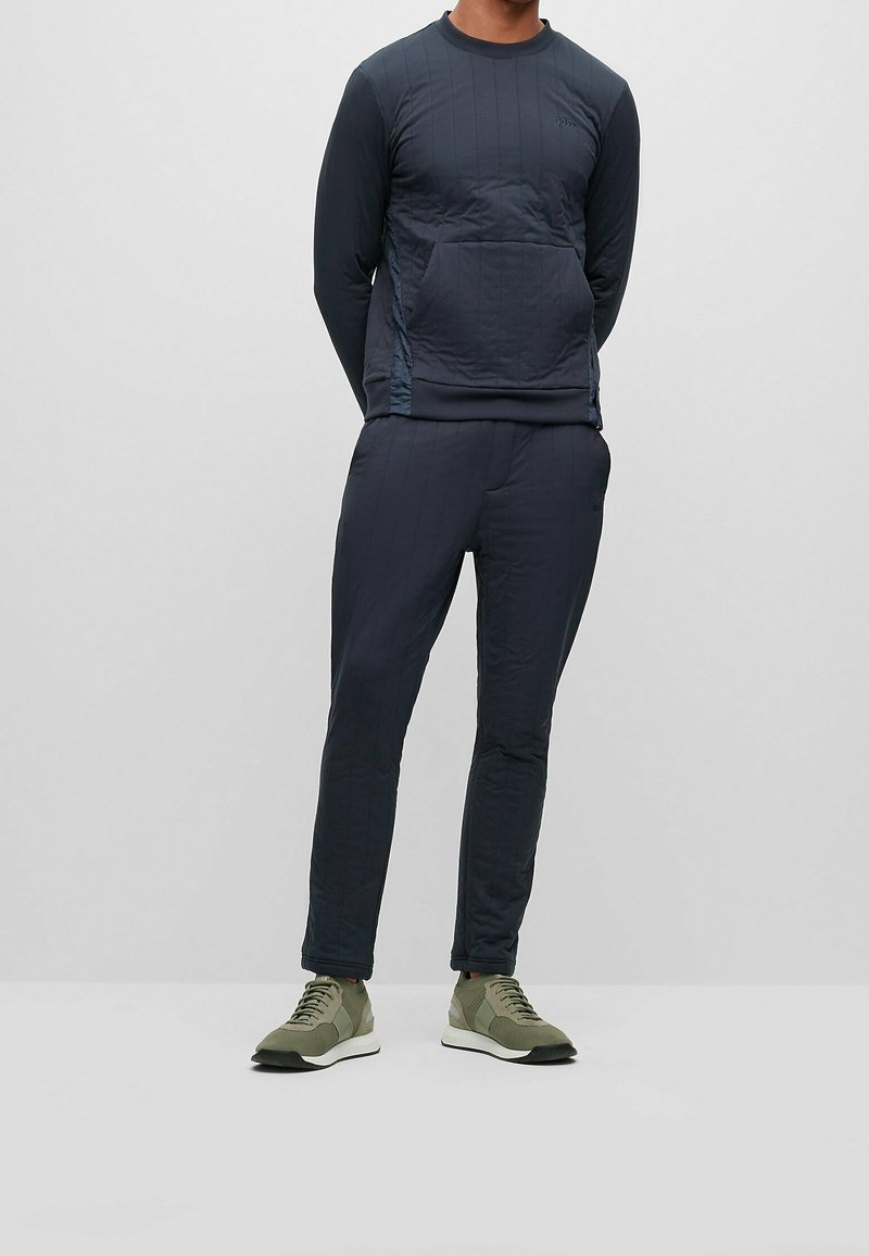 Homme debout portant un sweat-shirt à manches longues foncé avec une poche avant, un pantalon assorti et des baskets vertes sur un fond uni.
