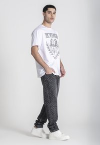 Biała bawełniana koszulka z czarnym grafiką, w połączeniu z ciemnymi, wzorzystymi dresami i białymi sneakerami. Stylizacja charakteryzuje się luźnym krojem.