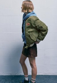 Giubbotto bomber verde oliva con una patch ricamata, indossato sopra una felpa con cappuccio azzurro chiaro, abbinato a una gonna in camoscio marrone e calzini a righe.