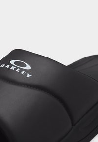 Μαύρο σανδάλι Oakley slide με φαρδιά λουρί που φέρει λευκό λογότυπο Oakley στην κορυφή.