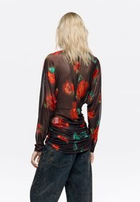Przezroczysta bluzka z długim rękawem w kwiatowy wzór w odcieniach czerwieni i zieleni na czarnym tle, marszczona w talii. Noszona z luźnymi spodniami dżinsowymi.