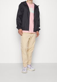 Svart lättviktsjacka, rosa grafisk t-shirt, beige byxor och grå sneakers med lila accenter. Avslappnat outfit med en lös passform.