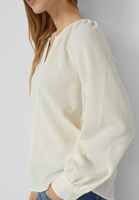 Blusa bianca testurizzata con un sottile motivo a pieghe, scollatura a V e dettaglio arricciato sulle spalle. Maniche lunghe con polsini aderenti.