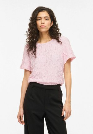Femme aux cheveux bouclés foncés portant un haut rose texturé à manches courtes et un pantalon noir taille haute, debout devant un fond uni.