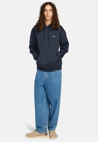 Felpa in cotone blu navy con tasca frontale, cappuccio con cordino e logo. Abbinata a jeans larghi azzurri e scarpe beige.