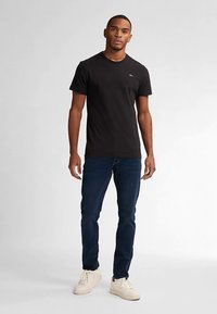 Czarna bawełniana koszulka z okrągłym dekoltem, w połączeniu z ciemnoniebieskimi jeansami i jasnymi trampkami, stojąca na neutralnym tle.