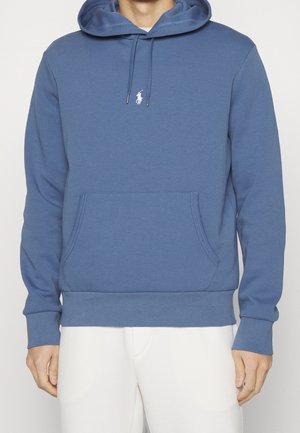 Man draagt een blauwe capuchontrui met voorzak en wit polo-spelerlogo, gecombineerd met witte broek, staand tegen een effen achtergrond.