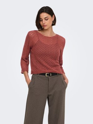 Femme aux cheveux courts et foncés portant un pull en maille couleur rouille et un pantalon large marron taille haute, les mains dans les poches.