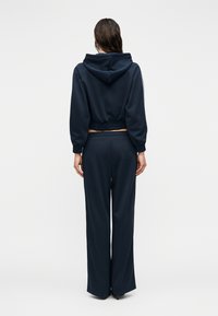 Γυναίκα που φοράει κοντό μπλε φούτερ με ελαστική μέση και εναρμονισμένο φαρδύ παντελόνι, εμφανιζόμενη από πίσω σε μονόχρωμο φόντο.