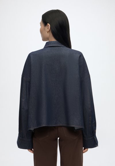 Femme aux longs cheveux foncés portant une veste en jean bleu foncé ample et un pantalon marron, tournée de dos sur un fond clair uni.