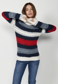 Un pull en tricot chunky multicolore présentant des rayures bleu marine, rouge, gris et crème, un col montant et un ourlet côtelé avec des accents en forme de diamant.