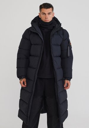 Uomo che indossa un lungo piumino nero con cappuccio, pantaloni neri e dolcevita nero, in piedi davanti a uno sfondo chiaro e uniforme.