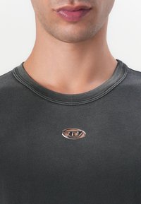 Tmavošedé tričko s krátkym rukávom a okrúhlym výstrihom, s metalickým oválnym logom v strede hrude. Hladká textúra látky.