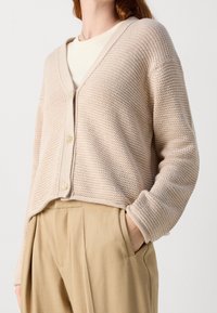 Cardigan beige lavorato a maglia con scollo a V, dotato di chiusura frontale con bottoni e polsini a costine, abbinato a pantaloni beige.