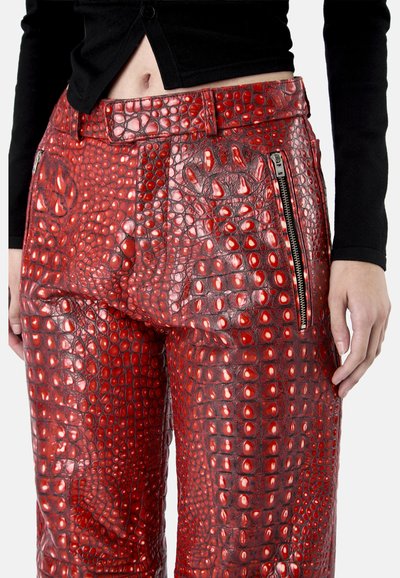 Personne portant un pantalon en cuir rouge verni effet crocodile taille haute avec une poche zippée à l'avant et un top court noir.