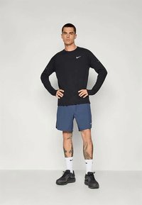 Mann steht mit den Händen in der Hüfte, trägt ein schwarzes Langarmshirt von Nike, blaue Shorts, weiße Nike-Socken, schwarze Sneakers und hat sichtbare Bein-Tattoos.
