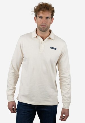 Polo de manga larga color beige claro con cuello de solapa, tres botones y una etiqueta azul que dice "AMARO JEANS" en el pecho izquierdo.