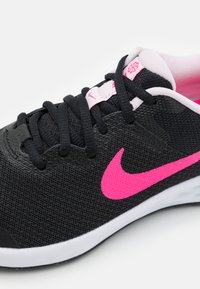 Zapatilla deportiva negra con parte superior de malla texturizada, logo de Nike rosa, cordones negros y detalles rosas en el cuello y el interior. Suela blanca.