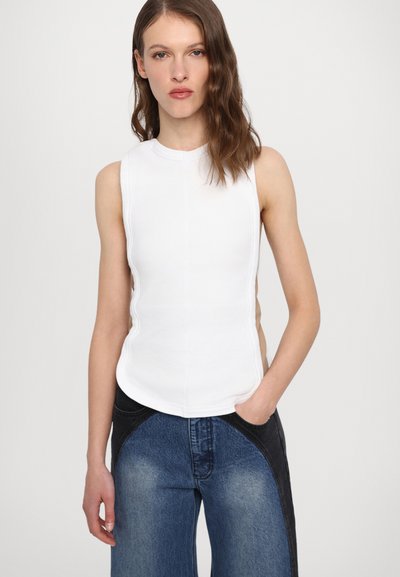 White sleeveless top with ribbed texture, curved hem, and side cutouts, paired with blue denim wide-leg pants featuring black accents.