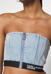 Nainen, jolla on yllään olkaimeton vaaleansininen farkkCrop-top, jossa on edessä vetoketju ja musta nauha, jossa on teksti "Karl Lagerfeld Jeans."