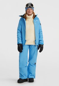Blauwe geïsoleerde skijas met een fleece kraag, gecombineerd met bijpassende blauwe broeken. De outfit omvat zwarte handschoenen en een skihelm.