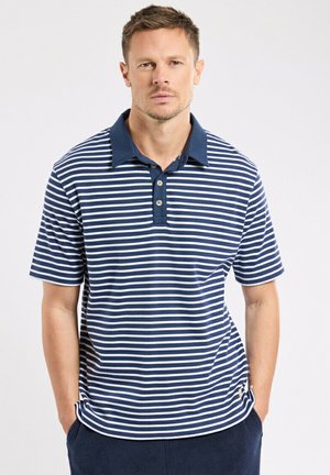 Homme portant un polo à manches courtes à rayures horizontales bleu marine et blanches avec un col bleu marine, debout les mains dans les poches.