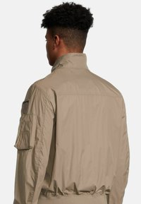 Beige lichtgewicht jasje met een hoge kraag, elastische tailleband en een schouderzak. Glad textuur met subtiele glans.