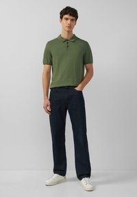 Zielona koszulka polo z krótkim rękawem i trzema guzikami, połączona z ciemnym dżinsami i białymi trampkami, stojąca na neutralnym tle.