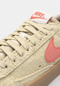 Nike sneaker, texturált bézs felsőrésszel, rózsaszín swoosh-sal és bézs fűzőkkel. Fehér logo található a nyelvén, és gumitalpa van.