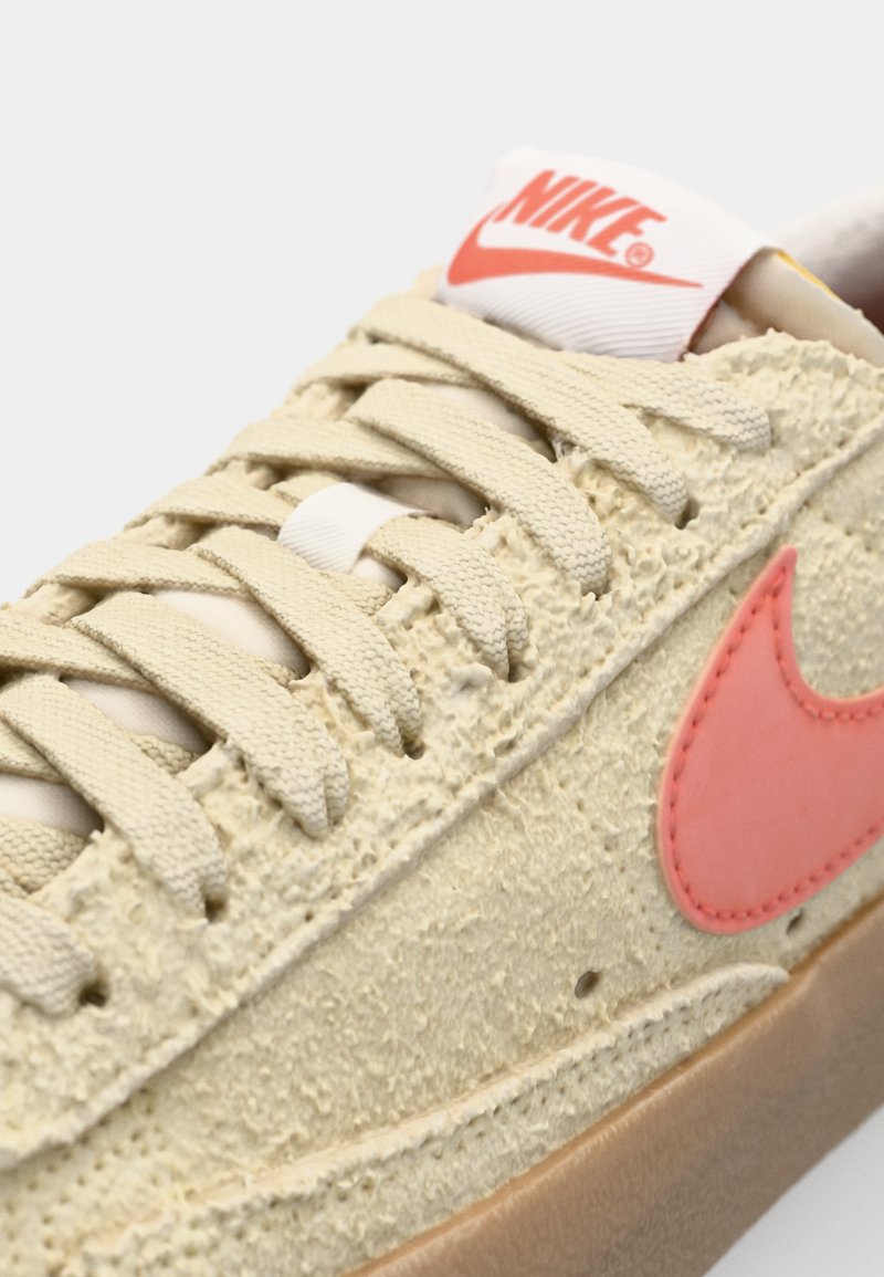 Baskets Nike avec un dessus beige texturé, un swoosh rose et des lacets beiges. Comprend un logo blanc sur la languette et une semelle en gomme.