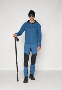 Chaqueta deportiva azul con cremallera frontal y dos bolsillos, combinada con pantalones bicolor en azul y negro. El modelo sostiene un bastón de senderismo.
