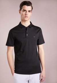 Svart polo skjorte laget av myk bomull. Har en klassisk krage, tre knapper i placket, og en liten logo på venstre bryst. Korte ermer.