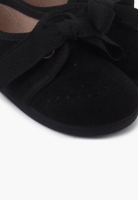 Pantofi din suede negru cu talpă plată, prezentând o panglică lată neagră legată sub formă de fundă în față. Textură netedă, cu perforații decorative.