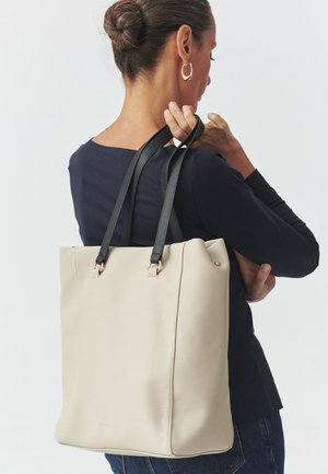 Beige tekstureret læder tote taske med sorte læderhåndtag og guld hardware-accenter. Simpel, struktureret design med et rummeligt interiør.