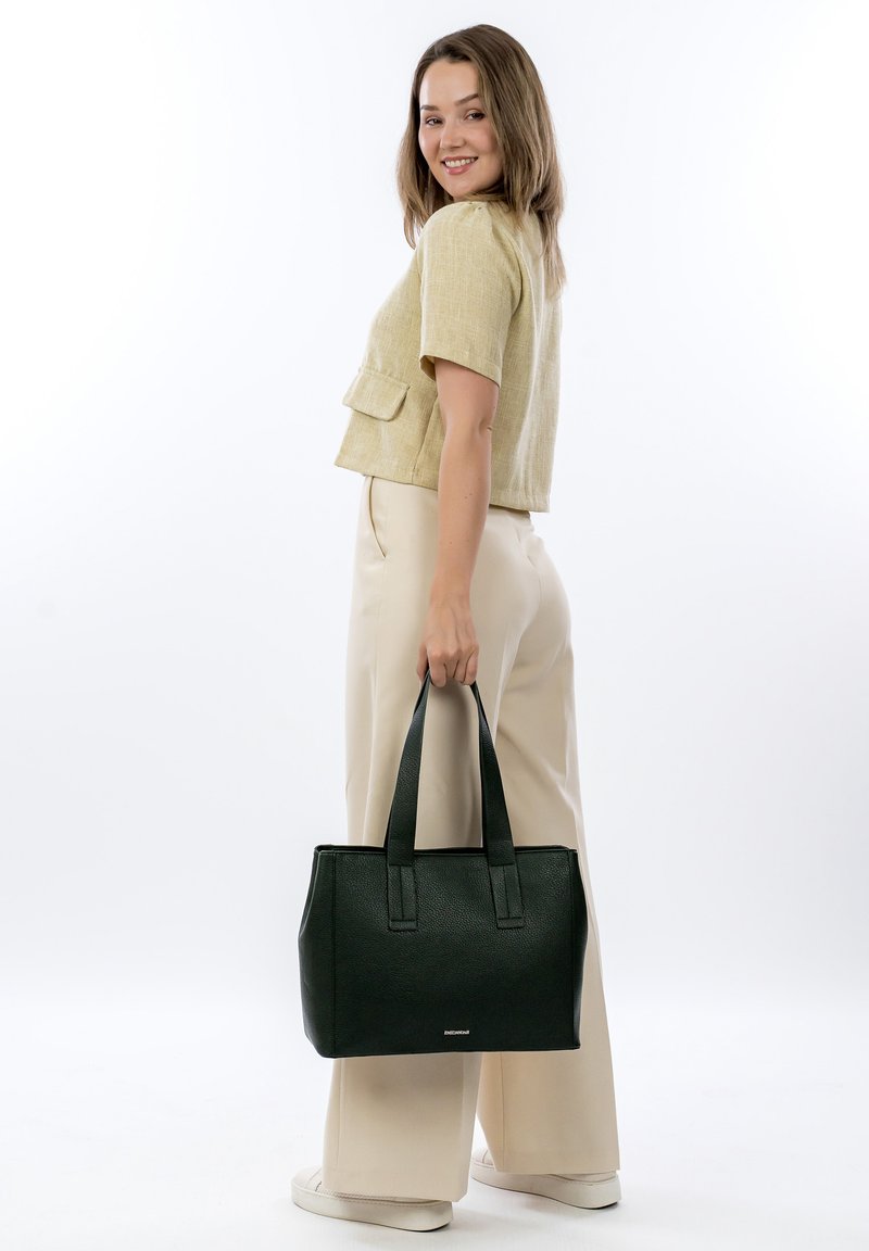 Borsa nera con trama testurizzata, manici doppi, forma strutturata e dettaglio del logo, abbinata a una blusa chiara a maniche corte e pantaloni beige.
