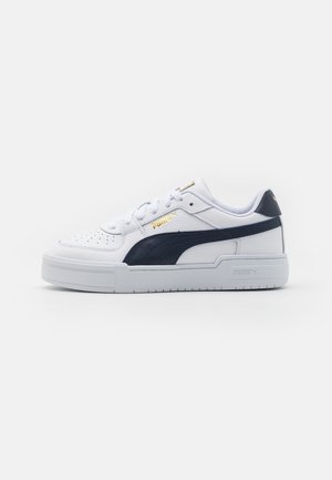 Biały niski sneaker z czarnym paskiem po boku, złotym logo Puma, perforowanym noskiem i grubą podeszwą na jednolitym białym tle.