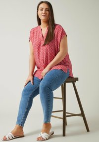 Blusa de patrón rojo con mangas cortas, combinada con unos jeans ajustados azules y sandalias blancas. La blusa presenta un diseño de zigzag y un cierre de botones en el frente.