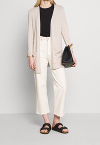 Béžový kardigan s kapsami, černá košile, bílé široké kalhoty a černé sandály. Taška má černý trojúhelníkový tvar s rukojetí.