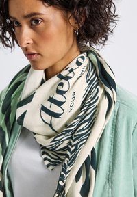 Ung kvinna med lockigt hår som bär en vit skjorta, ljusgrön jacka och en krämfärgad scarf med svart text och zebramönster.