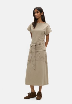 Kobieta stoi, ubrana w beżową bluzkę z krótkim rękawem i pasującą do niej midi spódnicę z haftem, w połączeniu z brązowymi zamszowymi botkami z frędzlami przy kostce.