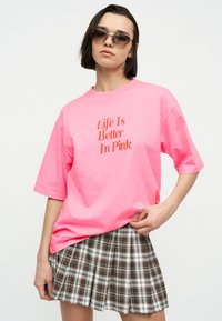 Γυναίκα με φιμέ γυαλιά ηλίου, φωτεινό ροζ υπερμεγέθες μπλουζάκι με το κείμενο «Η Ζωή Είναι Καλύτερη σε Ροζ» και καφέ καρώ πλισέ φούστα.