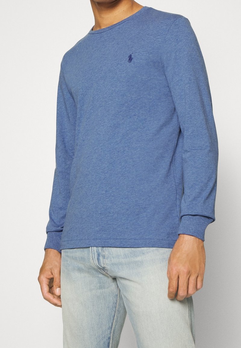 Långärmad blå bomullsskjorta med rund hals, med en liten mörk logga på bröstet och ribbade ärmslut. Bärs med ljusa denimjeans.