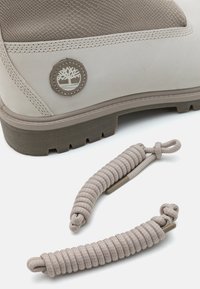 Stivale impermeabile beige con parte superiore texturizzata, patch logo marrone e suola robusta. Include lacci beige abbinati con design a spirale.