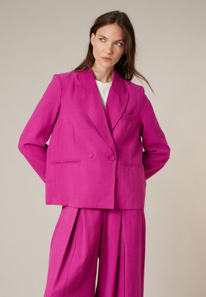 Woman with long brown hair wearing a bright pink double-breasted blazer and matching wide-leg trousers looking to the side.