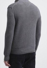 Uomo che indossa un maglione grigio a maglia aderente con polsini e orlo a coste, visto da dietro su uno sfondo chiaro e semplice.