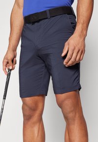 Marineblå shorts laget av lett materiale med flat front, sidelommer og et svart belte med et logo. En golfklubbe holdes i den ene hånden.
