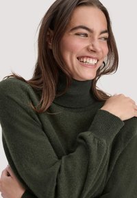 Dunkelgrüner Rollkragenpullover, weiche Textur, lockere Passform, gerippte Bündchen und hoher Kragen. Model lächelt mit leicht zerzausten Haaren.